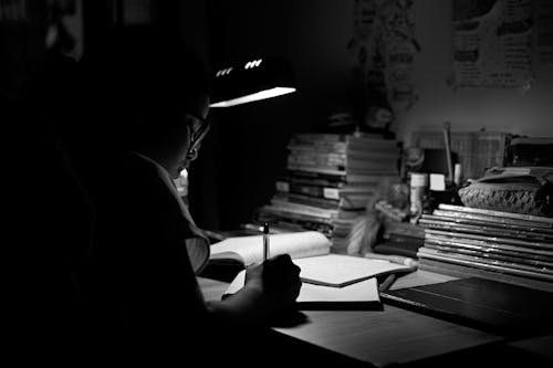 Free A student intensely studying under a desk lamp, surrounded by books, in a black and white setting. Stock Photo